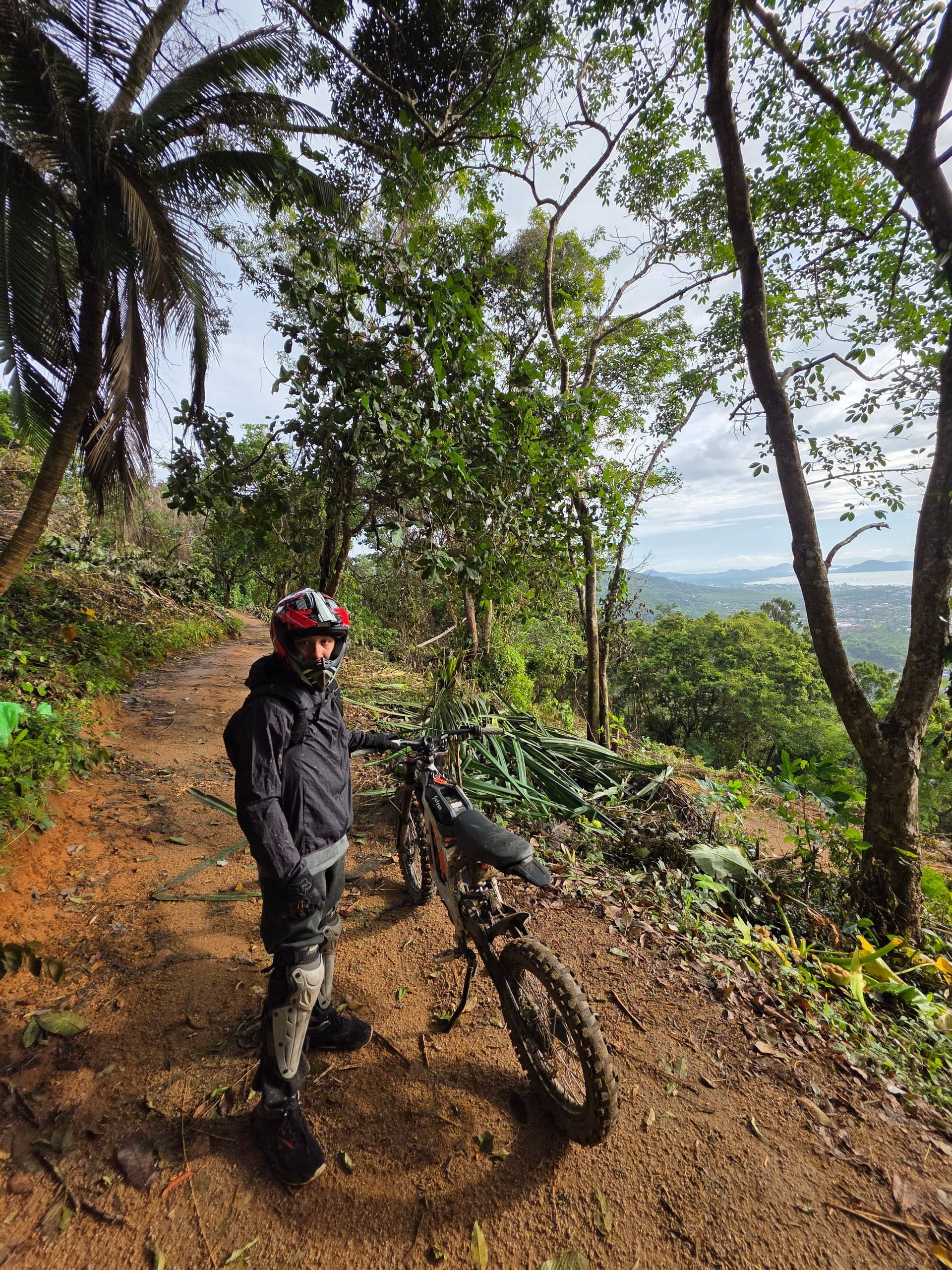 A person with a Surron electric motorbike on a dirt trail surrounded by lush greenery in Phuket, Thailand.